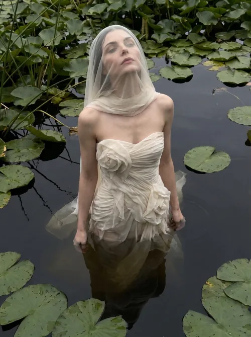 Made on Blogent: ethereal fine-art fashion portrait, a woman in a white draped gown standing waist-deep in a lily pond, soft overcast…
