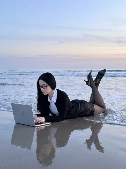 Made on Blogent: cinematic lifestyle shot, woman in a black blazer working on a laptop while lying on a wet beach at sunset, mirror…