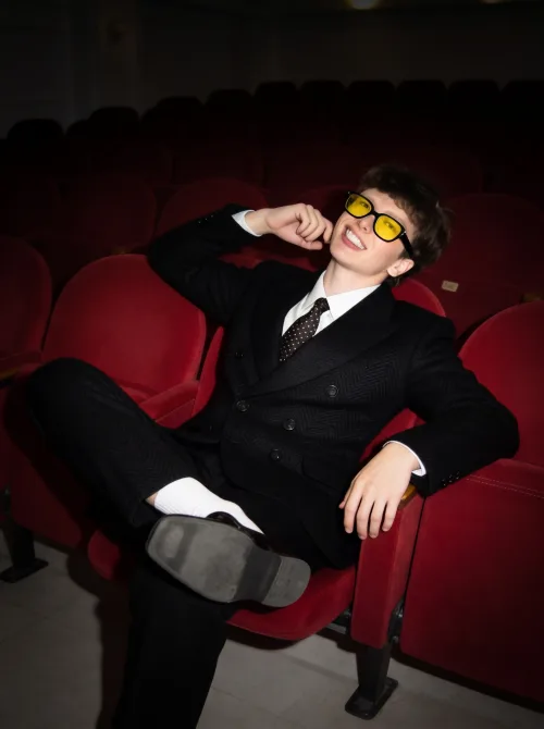 Made on Blogent: high-fashion editorial, model in a sharp black suit reclining in a red velvet theatre seat, tinted sunglasses…